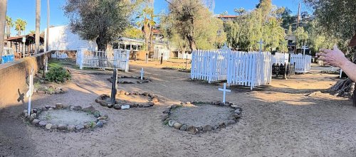 A sandy area with several circular plots marked by stones and white crosses, surrounded by trees and a white fence.