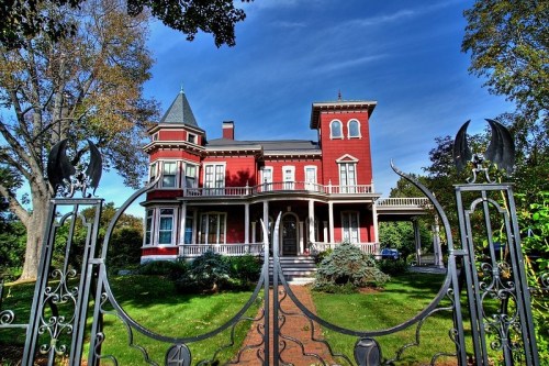 A grand red Victorian house with a turret, surrounded by greenery and a decorative wrought-iron gate under a blue sky.
