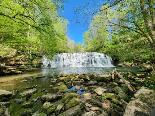 A serene waterfall cascades over rocks, surrounded by lush green trees and a clear blue sky.