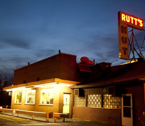 A retro diner named Rutt's Hut illuminated at dusk, featuring a neon sign and a warm glow from the windows.
