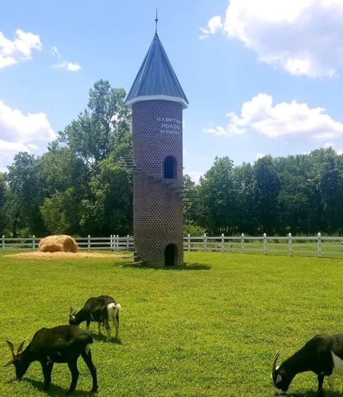 A brick tower with a pointed roof stands in a grassy field, with goats grazing nearby and trees in the background.