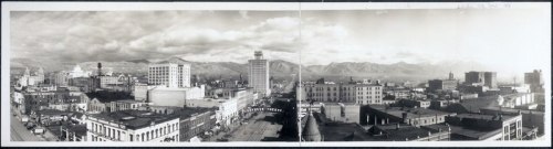 Panoramic view of a city skyline with mountains in the background, featuring buildings and streets under a cloudy sky.