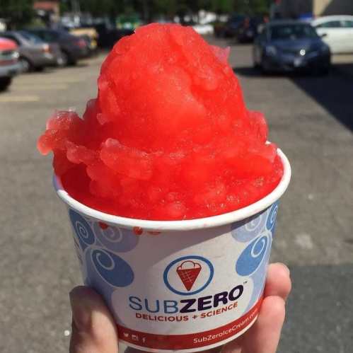 A hand holding a cup of bright red snow cone from SubZero, with a sunny parking lot in the background.