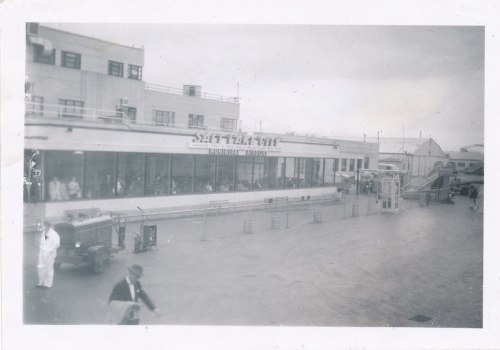 Black and white photo of a busy waterfront building with people, vintage cars, and a cloudy sky.