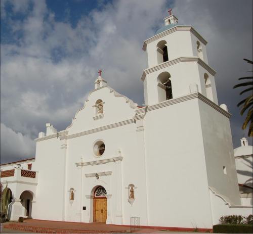 A white church with a bell tower, featuring decorative elements and a wooden door, set against a cloudy sky.