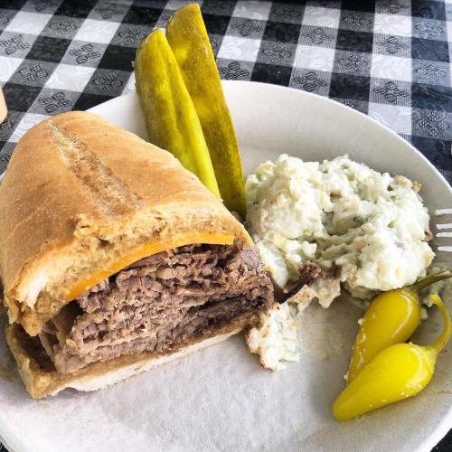 A sandwich with beef and cheese, served with pickles, peppers, and a side of potato salad on a paper plate.