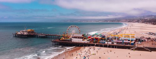 Aerial view of a vibrant beach pier with a Ferris wheel, surrounded by sandy shores and ocean waves under a cloudy sky.