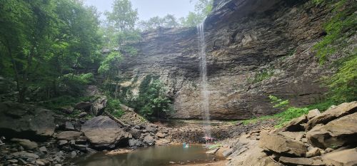 A serene waterfall cascades down a rocky cliff into a calm pool, surrounded by lush greenery and large boulders.