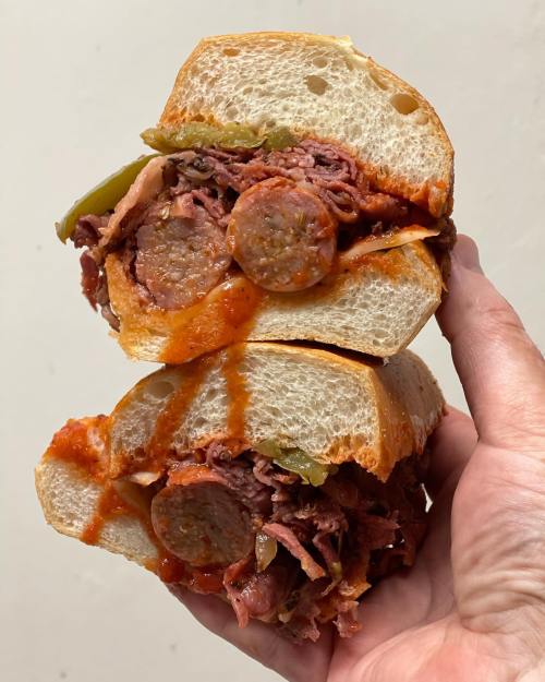 A hand holding a stacked sandwich filled with sausage, beef, peppers, and marinara sauce on a soft roll.