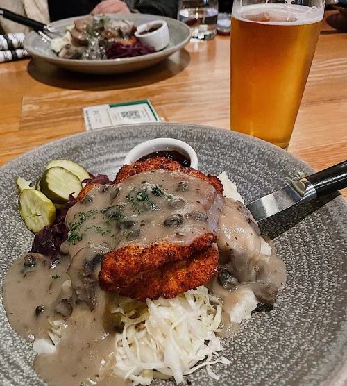 A plate of fried chicken topped with gravy, served with pickles and cabbage, alongside a glass of beer.