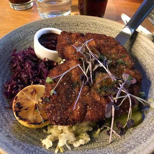 A plate featuring two breaded cutlets, pickles, red cabbage, a lemon slice, and a small bowl of sauce.