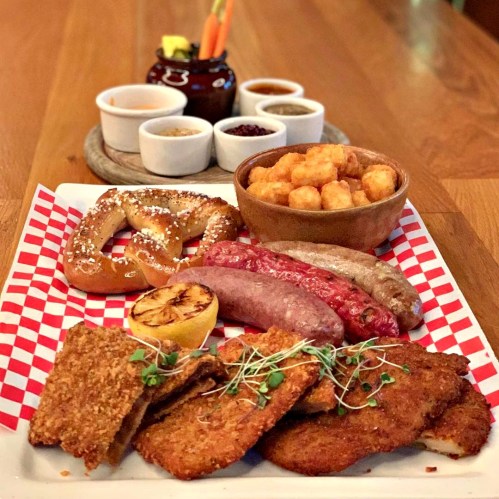 A platter featuring pretzel, sausages, chicken, tater tots, and dipping sauces on a wooden table.