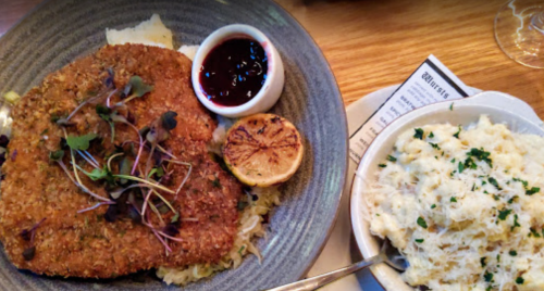 A plate featuring a breaded cutlet with sauce, grilled lemon, and a side of creamy mashed potatoes, garnished with herbs.