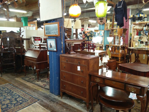 A cozy antique shop filled with wooden furniture, including dressers, tables, and lamps, set in a rustic environment.
