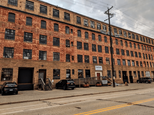 A large, weathered brick building with multiple windows and a sign, alongside parked cars and a street.