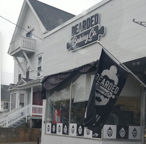 Exterior of Bearded Baking Co. with a flag and a white building in a rainy setting.