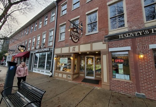 A street view of a brick building with shops, including a bakery, and a person walking by on a rainy day.