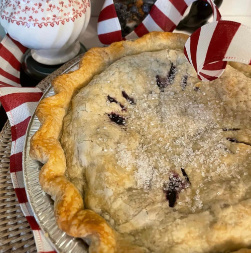 A close-up of a homemade pie with a golden crust, sprinkled with sugar, surrounded by festive red and white ribbons.