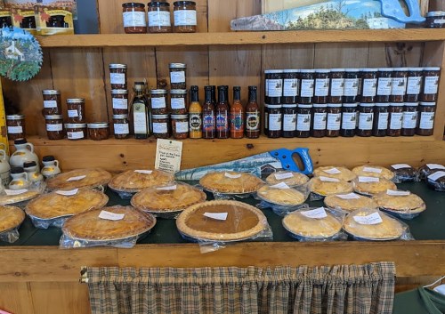 A wooden shelf displays various pies and jars of preserves, with a saw and a checkered cloth in the foreground.
