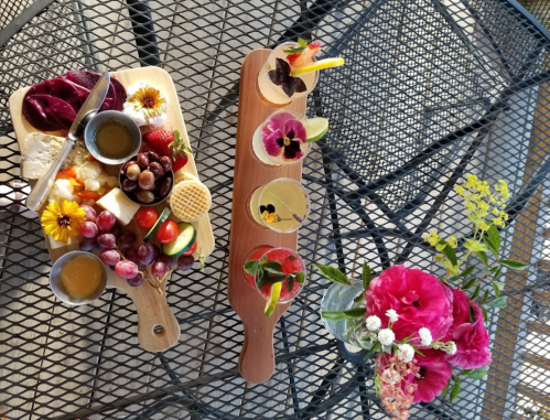 A vibrant spread of cheese, fruits, and drinks on a table, accompanied by colorful flowers.
