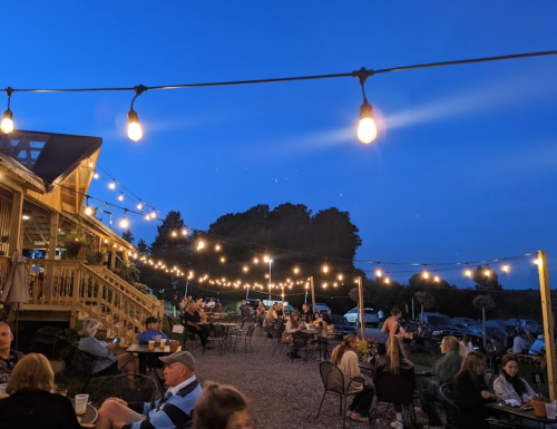 Outdoor dining area at dusk, with string lights illuminating tables and patrons enjoying the evening atmosphere.