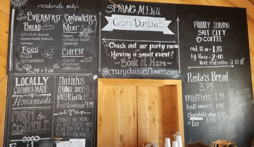 Chalkboard menu featuring breakfast sandwiches, drinks, and local offerings at Crazy Daisies, with event booking info.