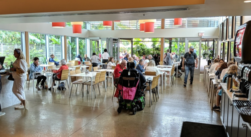 A busy restaurant with large windows, filled with diners and staff, surrounded by greenery and bright orange light fixtures.
