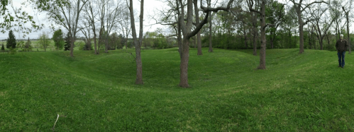 A panoramic view of a grassy landscape with trees and gentle hills, featuring a person walking in the distance.