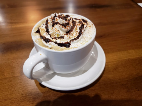 A white cup of coffee topped with whipped cream, chocolate drizzle, and a sprinkle of cocoa, sitting on a wooden table.