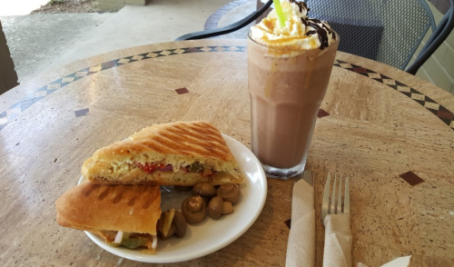 A toasted sandwich with vegetables and olives, alongside a tall glass of chocolate milkshake topped with whipped cream.