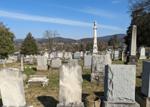 A cemetery with various gravestones and a tall monument, set against a backdrop of rolling hills and a clear blue sky.