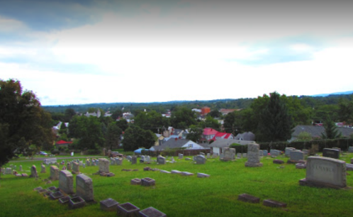 A cemetery on a hillside overlooking a small town with houses and trees under a cloudy sky.