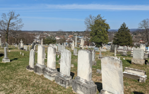A cemetery with weathered gravestones, green grass, and a view of a town in the background under a clear blue sky.