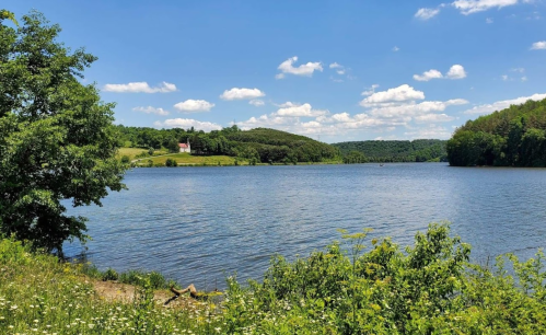 A serene lake surrounded by green hills and a blue sky with fluffy clouds, featuring a small house in the distance.