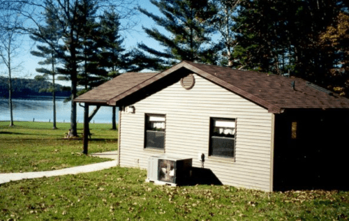 A small brown cabin by a lake, surrounded by trees and a grassy area, with a pathway leading to the entrance.