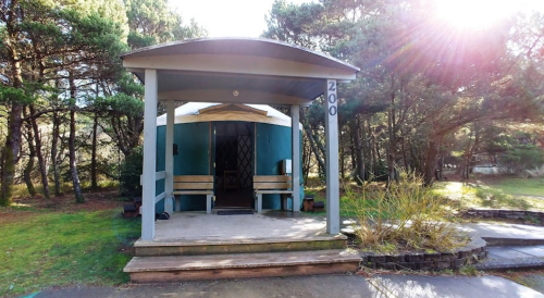 A small blue cabin with a porch, surrounded by trees and sunlight, set in a grassy area.
