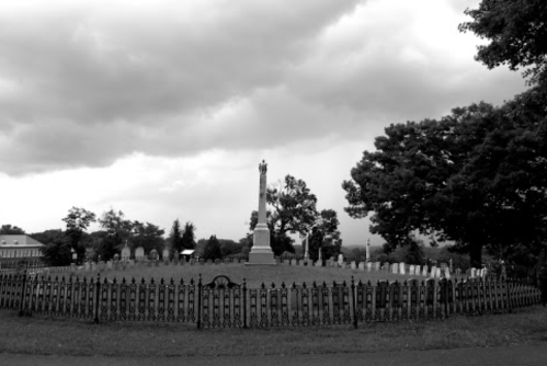 A black and white image of a cemetery with a tall monument surrounded by a wrought iron fence and cloudy skies.