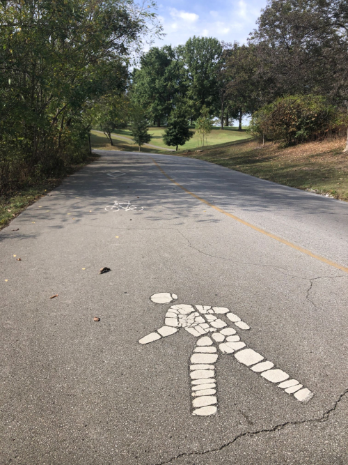 A paved path with a stick figure painted on it, surrounded by trees and greenery in the background.