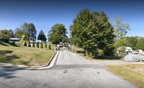 A tree-lined road leads up a gentle slope, with houses visible in the background and a few people walking ahead.