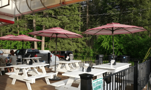 Outdoor dining area with white picnic tables and red umbrellas, surrounded by trees.