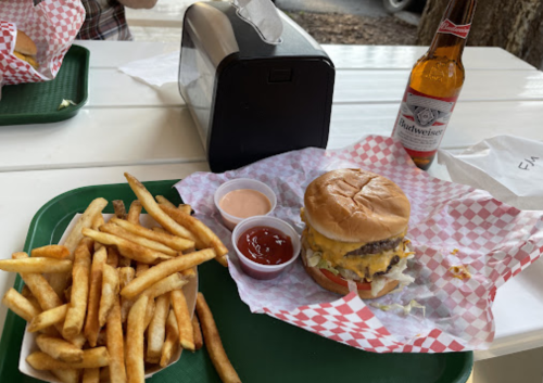 A tray with a cheeseburger, fries, dipping sauces, and a beer bottle on a picnic table.