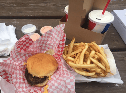 A burger in a red checkered wrapper, served with fries and two drinks in takeout containers on a wooden table.
