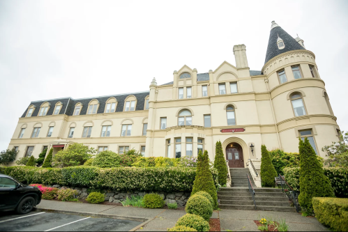 A large, elegant building with a turret, surrounded by greenery and flowers, under a cloudy sky.