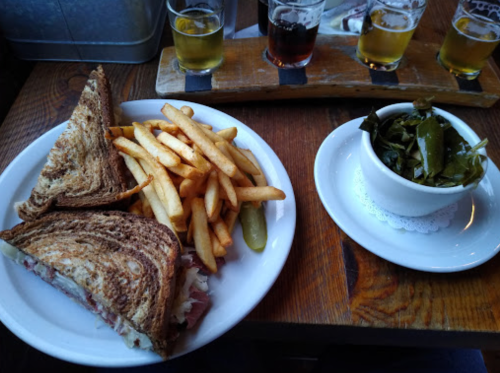 A plate with a sandwich, fries, a pickle, and a small bowl of greens on a wooden table.