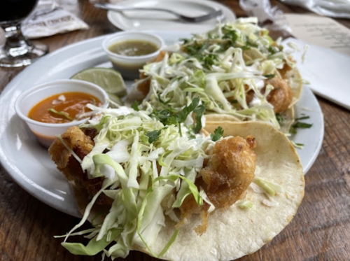 Two crispy fish tacos topped with shredded cabbage and cilantro, served with lime and two dipping sauces on a plate.