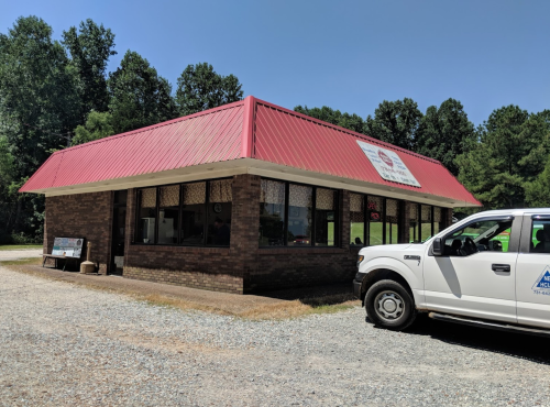 A brick building with a red roof and large windows, parked white truck in front, surrounded by trees and gravel.