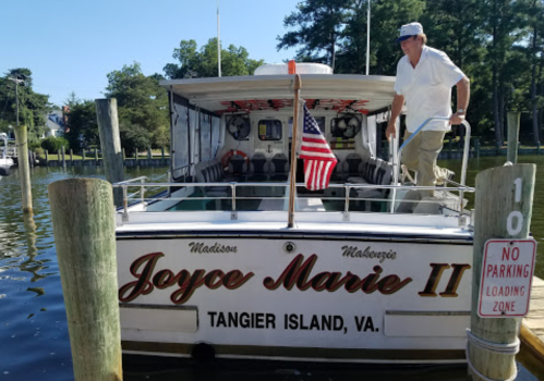 A man stands on the deck of the "Joyce Marie II" boat, docked at Tangier Island, VA, with an American flag displayed.