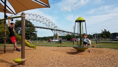 Children play on a playground with a slide and swing, with a bridge and cars in the background under a blue sky.