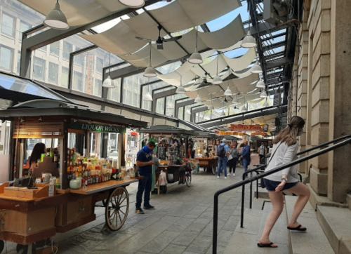 A bustling market with vendors, shoppers, and shaded stalls under a glass roof, featuring various goods and food options.