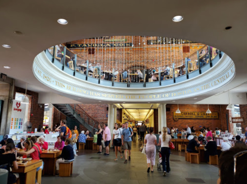 A bustling indoor market with people dining and shopping, featuring a circular design and brick walls.